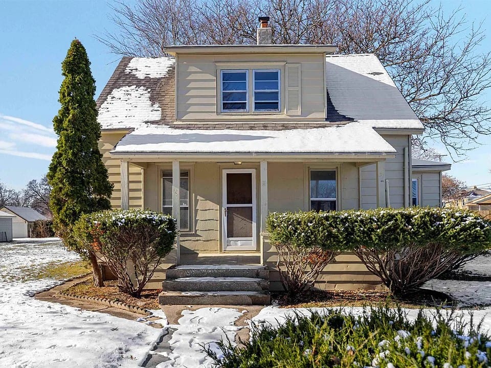 A cream-sided storey-and-a-half house with a wide shed dormer and a full-width covered front porch, photographed after snowfall.