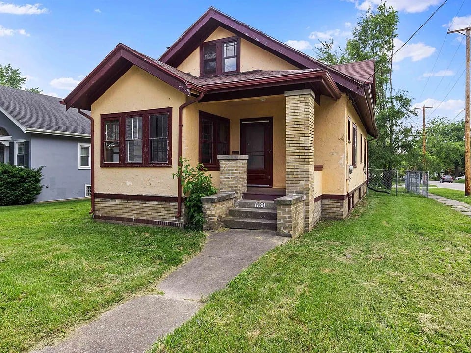 A yellow stucco craftsman bungalow with maroon trim, a gabled front porch on brick piers, and the number 638 cast into the front step.